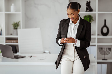 Poster - Happy black female entrepreneur in formal wear reading message on smartphone, standing at office workplace table
