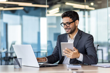 Young successful hispanic businessman at workplace using app on tablet computer, smiling man inside office building, working with laptop, wearing business suit, happy with achievement.