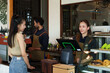 © Ekkasit A Siam - Young female shopkeeper wearing an apron Taking orders from woman customers in front of the counter and her husband is serving as a barista Atmosphere in a coffee shop, a small family business..
