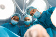 Multiethnic medical team of surgeons performing an operation wearing operating gowns, surgical caps, masks and gloves while closely looking at patient with fully concentration from above.