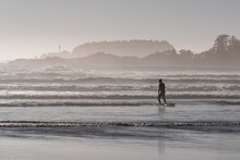 Unrecognizable Surfer Silhouette In The Strong Waves On Cox Bay Beach Of Tofino With Lighthouse In Background, Tofino, Vancouver Island, British Columbia, Canada.