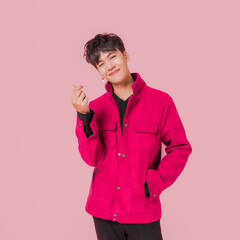 Portrait of a happy Asian handsome man in fashionable pink jacket clothing standing smiling and using fingers to make a mini heart sign isolated on pink background.