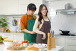 © Bangkok Click Studio - Asian young lovely lover couple husband and wife in casual outfit standing smiling male helping wearing clean striped apron to female in full decorated modern kitchen with cooking equipment at home