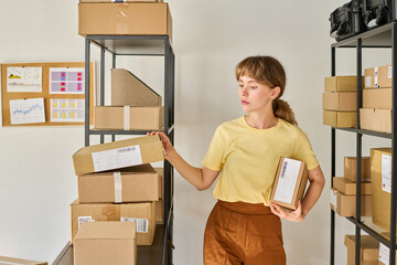 Young female worker of marketplace or storage room standing by rack with packed parcels and looking for box with necessary number