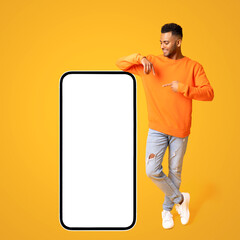 Optimistic happy indian guy posing by huge smartphone with empty blank screen, pointing at display and smiling, studio shot isolated on yellow, mockup