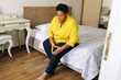 Leinwandbild Motiv African american aged plus size female in yellow shirt and glasses sitting on bed in her bedroom feeling pain in her left knee, suffering from bone disease, touching and massaging leg with hands