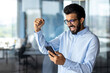 © Tetiana - Indian young man working in an office center, standing and using the phone. Looks at the screen and happy shows a victory gesture with his hand