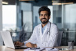 Portrait of a young doctor of Indian origin working in the office using a laptop, sitting at the table and smiling at the camera