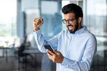 Indian young man working in an office center, standing and using the phone. Looks at the screen and happy shows a victory gesture with his hand
