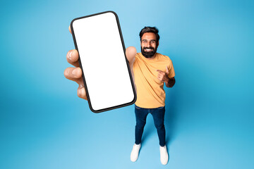 Cheerful indian man holding cellphone with white empty screen, pointing at blank display for advertisement, mockup