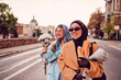 © Minet - Couple woman one wearing a hijab and a modern yet traditional dress, and the other in a blue dress and scarf, walking together through the city at sunset. One carries a bouquet and bread, while the