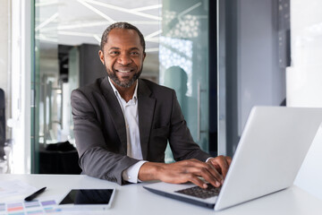 Portrait of successful happy african american boss, man smiling and looking at camera, businessman in business suit sitting at desk with laptop inside office.