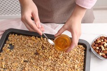 Making Granola. Woman Adding Honey Onto Baking Tray With Mixture Of Oat Flakes And Other Ingredients At White Table In Kitchen, Closeup