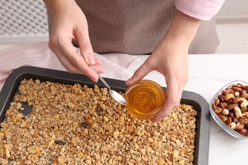 Making granola. Woman adding honey onto baking tray with mixture of oat flakes and other ingredients at white table in kitchen, closeup