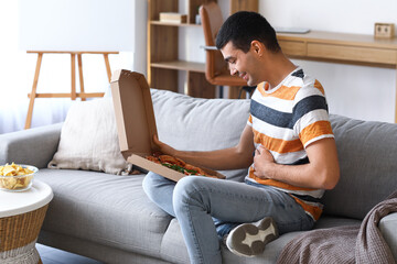 Wall Mural - Young man with box of tasty pizza sitting on sofa at home