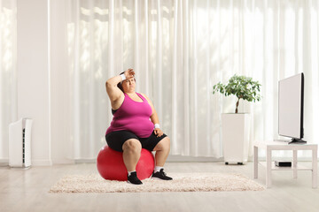 Poster - Tired overweight woman sitting on a fintess ball in front of tv