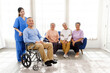 © artitwpd - The caregiver therapist stands with an Asian senior sitting in a wheelchair with a group of senior women sitting on a sofa in the background. The nursing home facilitates a support group.