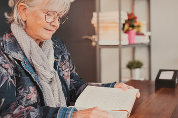 Senior woman sitting at cafe table reading a book. Quiet retirement lifestyle for an elderly caucasian female in eyeglasses and denim jacket