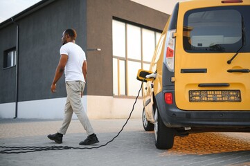 Wall Mural - A african american man stands next to yellow electric delivery van at electric vehicle charging stations