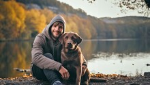 A Young Guy With His Chocolate Labrador Dog Sits Hugging On The Shore Of A Mountain Lake On An Autumn Evening.