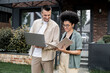 © LIGHTFIELD STUDIOS - smiling multicultural property agents standing with laptop and folder near modern city cottage