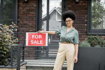 Wall Mural - optimistic african american property agent standing with for sale signboard near fence of cottage