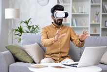 Surprised Serious Man In Virtual Reality Glasses Reviewing Course In Virtual Meeting Sitting On Sofa In Living Room At Home