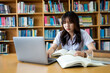 © EduLife Photos - Young Asian female university student concentrate on studying, doing assuagement project in library with books and laptop on table