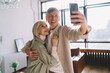 © BullRun - Cheerful senior woman and man taking selfie on mobile phone in apartment