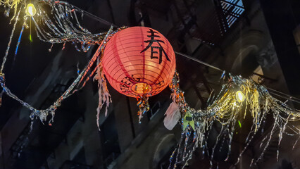 A red lantern with the Chinese character saying spring hanging among the street lights. There are some other decorations around the lights. Celebrating Chinese Spring Festival. City at night