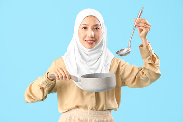 Poster - Young Asian woman in hijab with saucepan and ladle on blue background