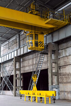 Yellow Overhead Crane With Linear Traverse And Hooks In Engineering Plant Workshop. Cabin Of Crane Operator And Landing Staircase.