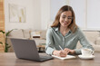 © New Africa - Happy woman writing something in notebook near laptop at wooden table in room