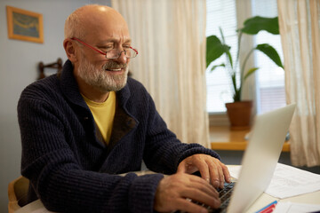 Side view of senior male professor in glasses and cardigan working on laptop sitting at table next to window at home, teaching online. Elderly man with beard chatting wit his friends on computer