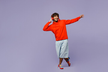 Full body smiling fun young Indian man wear red orange hoody casual clothes listen to music in headphones dance isolated on plain pastel light purple color background studio portrait Lifestyle concept