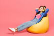 © ViDi Studio - Full body young woman of African American ethnicity she wear blue shirt casual clothes sit in bag chair listen to music in headphones isolated on plain pastel pink background studio Lifestyle concept