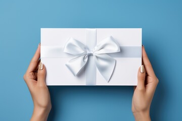 Wall Mural - Woman holding white present box with vivid silver ribbon bow as presenf for Christmas, fathers day, birthday on light blue background. Flat lay, copy space