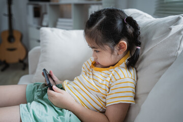 Wall Mural - Asian child girl looking using and touch mobile phone screen on couch sofa. Baby smiling funny time to use mobile phone. Too much screen time. Cute girl watching videos while tv, Internet addiction.