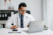 © Nuttapong punna - Confident Young Asian male doctor in white medical uniform sit at desk working on computer. Smiling use laptop write in medical journal in clinic.