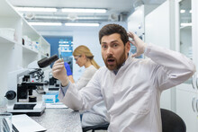 Portrait Of A Shocked Young Male Scientist Sitting In A Laboratory Behind A Microscope, Doing Research And Making A Discovery. He Holds The Substance In His Hand And Is Surprised By The Result.