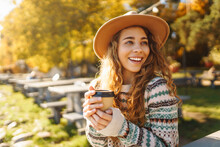 Portrait Of A Cute Woman In A Hat Sits With A Cup Of Coffee Overlooking The Lake. Outdoor Photo Of Relaxed Curly Woman Enjoying Hot Drink In Autumn Park.