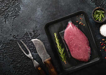 Wall Mural - Beef raw fillet steak in vacuum tray with pepper,salt and rosemary on black background with barbeque fork and knife.