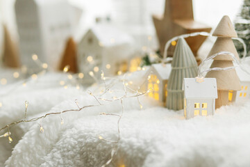 Wall Mural - Cozy christmas miniature village. Stylish cute little ceramic houses and wooden trees on soft snow blanket with glowing lights in room. Atmospheric winter village still life. Merry Christmas!