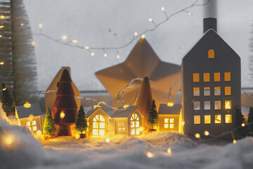 Cozy christmas miniature village. Stylish little ceramic houses and wooden trees on soft snow blanket with glowing lights in evening. Atmospheric winter village still life. Merry Christmas!