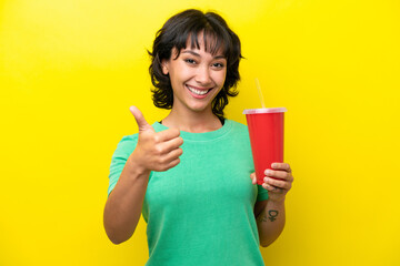 Wall Mural - Young Argentinian woman holding a soda isolated on yellow background with thumbs up because something good has happened