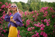 Happy smiling blonde woman wearing pale purple sunglasses, chunky chain, violet blouse, yellow trousers, holding mango flavor ice cream, posing in flower garden. Copy, empty space for text