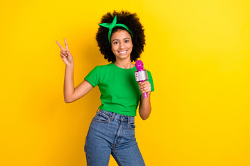 Poster - Photo of beautiful teen schoolgirl hold microphone demonstrate v-sign empty space isolated on yellow color background