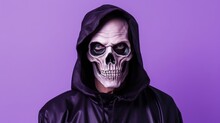 Man Dressed In Scary Halloween Costume. Studio Portrait Of Mr Death With Skeleton Skull Makeup On Face Wearing Black Cape With Hood Looking At Camera While Standing Isolated On Light Purple Background