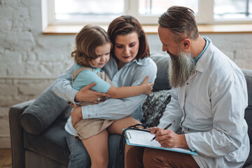 Doctor check heart during regular checkup in hospital. Sick little girl receive medical care. Senior male paediatrician examining sick kid sitting on mother's lap with stethoscope during home visit.