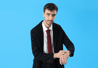 Wall Mural - Young man in suit looking at wristwatch on blue background